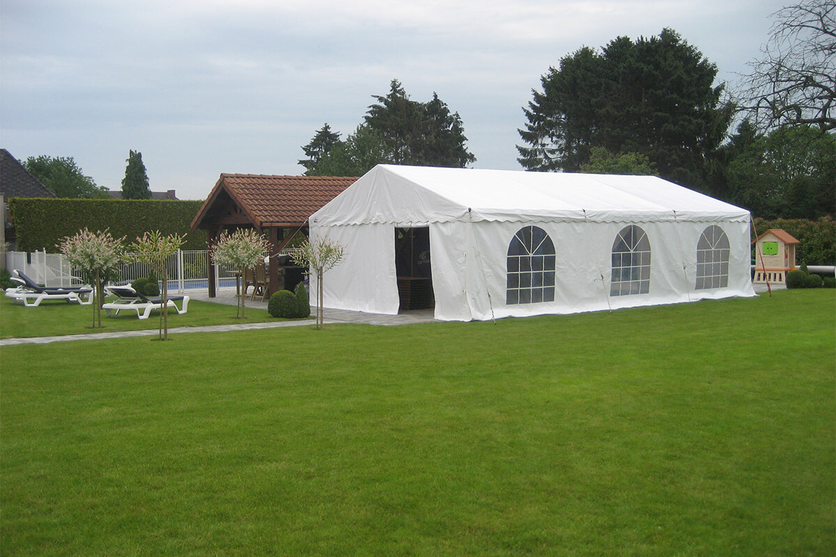 Vue d’une tente blanche installée dans un jardin bien entretenu, avec des transats et un abri en bois à proximité.